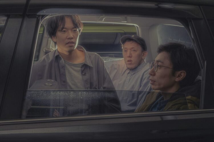 Photo of the three members of Kukangendai in a black car. The three men are dressed in casual clothing and sitting next to each other. One of them, sitting in the middle, is leaning forward and looking out of the right-hand window of the car. Another person is looking straight ahead, while the third person is looking at the person in the middle.