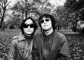 The two brothers of rock 'n' roll band SAHAJi are standing together in a park during the autumn period. There are leaves on the ground. Both musicians are facing the camera, wearing jackets and sunglasses. The photo is in black and white. Photography by Liz Gander
