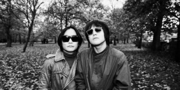 The two brothers of rock 'n' roll band SAHAJi are standing together in a park during the autumn period. There are leaves on the ground. Both musicians are facing the camera, wearing jackets and sunglasses. The photo is in black and white. Photography by Liz Gander