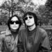 The two brothers of rock 'n' roll band SAHAJi are standing together in a park during the autumn period. There are leaves on the ground. Both musicians are facing the camera, wearing jackets and sunglasses. The photo is in black and white. Photography by Liz Gander