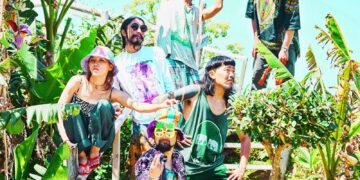 The photograph shows six members of Super Jet Kinoko striking various poses on a staircase, surrounded by green plants. All members are wearing colourful clothing, some complemented by colourful headwear and sunglasses.