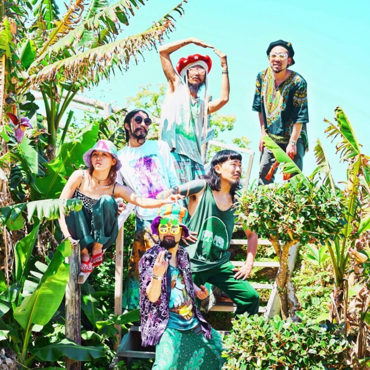 The photograph shows six members of Super Jet Kinoko striking various poses on a staircase, surrounded by green plants. All members are wearing colourful clothing, some complemented by colourful headwear and sunglasses.