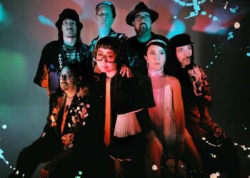 There are seven people in this photograph, all members of the psych-rock group TEKE::TEKE. They are standing close together, positioned against a light background and are wearing a mixture of dark and colourful clothing. Apart from the vocalist in the centre, no one is looking right into the camera. Photography by Flavie Lemée