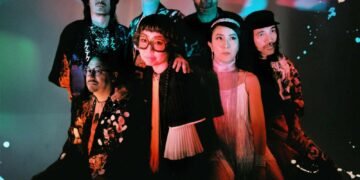 There are seven people in this photograph, all members of the psych-rock group TEKE::TEKE. They are standing close together, positioned against a light background and are wearing a mixture of dark and colourful clothing. Apart from the vocalist in the centre, no one is looking right into the camera. Photography by Flavie Lemée