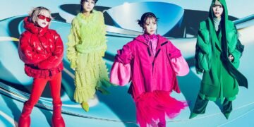 The four members of ExWHYZ in colourful outfits standing in a blue room with a futuristic looking background. Each member is dressed in a vibrant attire, all in a different colour.