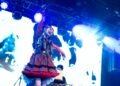 Photo of Utaha from Wednesday Campanella on stage, dressed in a red and blue outfit with tartan print. It shows an energetic scene where she looks toward the audience and waves her arms. Additionally, Kenmochi Hidefumi is seen in the background behind the DJ equipment. The stage is illuminated by white and blue lighting, also contributed by the big screen behind the two members. Photo by JJ Grant (Wonderlens) on behalf of AVO Magazine