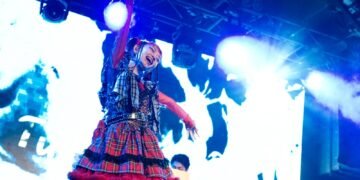 Photo of Utaha from Wednesday Campanella on stage, dressed in a red and blue outfit with tartan print. It shows an energe scene where she looks toward to audience and waves her arms. Additionally, Kenmochi Hidefumi is seen on the background behind DJ equipment. The stage is illuminated by white and blue lighting, also contributed by the big screen behind the two members.