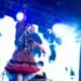Photo of Utaha from Wednesday Campanella on stage, dressed in a red and blue outfit with tartan print. It shows an energe scene where she looks toward to audience and waves her arms. Additionally, Kenmochi Hidefumi is seen on the background behind DJ equipment. The stage is illuminated by white and blue lighting, also contributed by the big screen behind the two members.