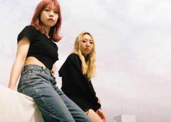 The photos features the two women of HYPER GAL sitting side by side on the roof of a building. Both are dressed in a black top and jeans, with their hair bleached or coloured, the two musicians look towards the camera lens. The photo is taken by Adam Armstrong (wespeakincolors in Instagram).