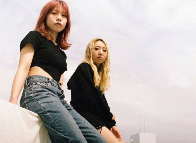 The photo features the two HYPER GAL women sitting side by side on the roof of a building. Both are dressed in a black top and jeans, with their hair bleached or coloured, the two musicians look towards the camera lens. The photo was taken by Adam Armstrong (wespeakincolors on Instagram).