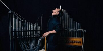 The photo shows a darkly lit room with sound artist and composer FUJI||||||||||TA standing in front of his hand-built pipe organ. The sound artist is dressed in black clothing and stands with his body turned to the right while his face is facing upwards and he appears to be screaming. Photo by Tomohiko Tagawa