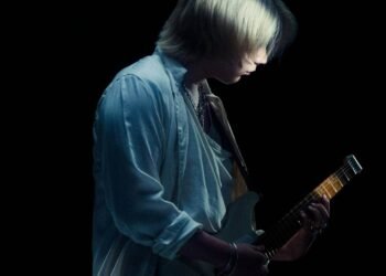 Photo captures Ichika Nito holding and playing his electric guitar in a dimly lit location, with a black background. It seems to be part of a photo shoot for his first album. © Ichika Nito