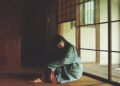 The photo depicts Ichiko Aoba sitting on a tatami mat in a dimly lit room as if she is lost in thought. She wears something with green and underneath red fabric. Photography by Kodai Kobayashi