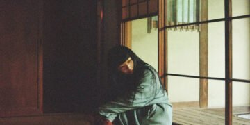 The photo depicts Ichiko Aoba sitting on a tatami mat in a dimly lit room as if she is lost in thought. She wears something with green and underneath red fabric. Photography by Kodai Kobayashi