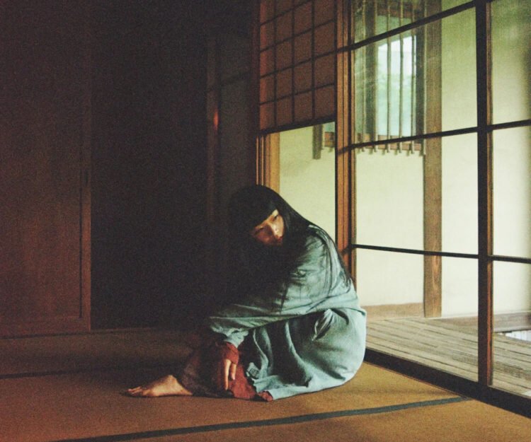 The photo depicts Ichiko Aoba sitting on a tatami mat in a dimly lit room as if she is lost in thought. She wears something with green and underneath red fabric. Photography by Kodai Kobayashi
