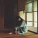 The photo depicts Ichiko Aoba sitting on a tatami mat in a dimly lit room as if she is lost in thought. She wears something with green and underneath red fabric. Photography by Kodai Kobayashi