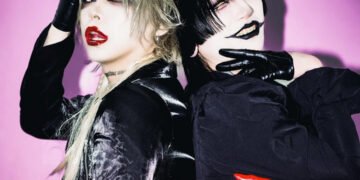 Photo of visual kei artists Aryu (THE DEVIL INSIDE) and Tsuzuku (tzkwym) standing next to each other with their backs to each other. They both look toward the camera. Both are fully dressed in black clothing, but with different garments and fabrics, wearing gloves, coloured lenses and topped off with full face of makeup. While Tsuzuku wears one red glove, Aryu wears red lipstick. © Killing Daze Management