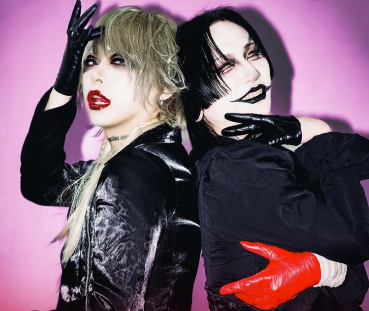 Photo of visual kei artists Aryu (THE DEVIL INSIDE) and Tsuzuku (tzkwym) standing next to each other with their backs to each other. They both look toward the camera. Both are fully dressed in black clothing, but with different garments and fabrics, wearing gloves, coloured lenses and topped off with full face of makeup. While Tsuzuku wears one red glove, Aryu wears red lipstick. © Killing Daze Management