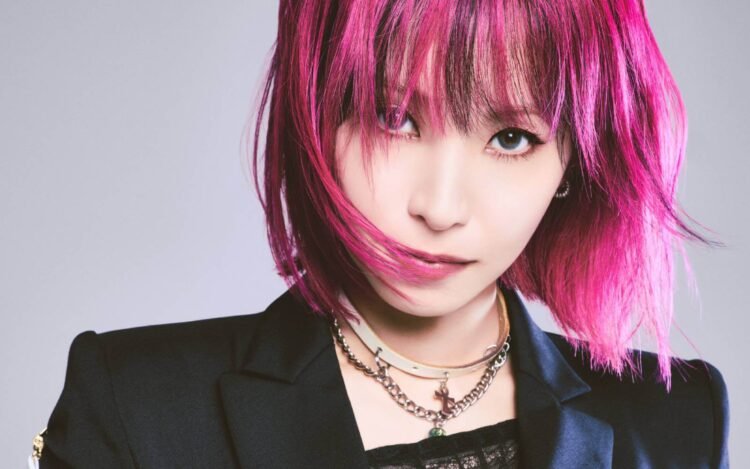 A cropped version of a vertical photo of LiSA with pink hair in a bob-style and a black jacket with a leather corset.