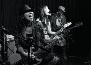 A black and white photograph that captured the energetic live performance of MIKABOMB at The Fox And Newt in Leeds. In the photo three musicians are visible, all playing their (bass) guitars. At the forefront is Mel Roxy with a head and Mika Handa on the right side, both facing the audience. Photography by JJ Grant (wonderlens) on behalf of AVO Magazine.