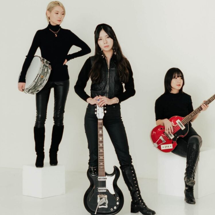 A group photo of The Highmarts as of 2025, used for the promotion of several performances in 2025, with the members Suzu, Minori and Shiori. They are all dressed in a black (leather) attire, while the background is white. Some members are posing on white blocks, all holding their instruments. © The Highmarts