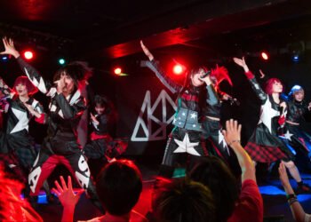 Live photo of the idol group ASP on stage, in the middle of performing a song. The stage is brightly lit with red and blue lights. They have their right arms raised while singing. The members are dressed in leather(-like) clothing, giving the idea that it is inspired by motor clothing. The clothing is mostly black but differently decorated, with red and blue stripes, white stars in different sizes, and other symbols or words. Some of them are fully dressed like that, while others have extra additions, like a tartan-style skirt. Photography by JJ Grant (wonderlens)