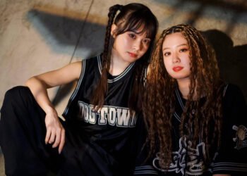 This photo shows two musicians from the duo EMNW sitting next to each other, Emma Yuki and Menu. They are sitting on what seems to be a skate ramp. The person on the left is wearing a black basketball jersey and has two braids in her hair. The person on the right has a black baseball jersey and has her head full with small braids and curly hair.