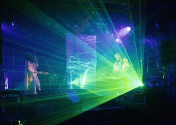 A live photo of Future Sound Lovers performing on a stage with lasers and LED lights. Kyra, the vocalist, is standing on the left and dressed in white clothing. Akira is standing on the right performing on his instrument behind a table.