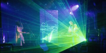 A live photo of Future Sound Lovers performing on a stage with lasers and LED lights. Kyra, the vocalist, is standing on the left and dressed in white clothing. Akira is standing on the right performing on his instrument behind a table.