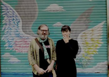 Jim O’Rourke and Eiko Ishibashi are standing together in front of a colourful closed shutter that is decorated with wings. They are positioned in such a way that it looks as though they both have wings. Eiko Ishibashi is dressed in black and is smoking, whilst Jim O’Rourke is wearing a black shirt, jeans, an ochre-yellow jacket and a green scarf with white dots. Photography by Mathieu Amalric
