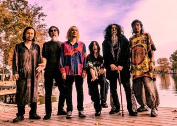 The photo shows six musicians standing next to each other on a deck near a lake. These  musicians are part of the music collective Acid Mothers Temple & The Melting Paraiso U.F.O. Most of the members are uniquely dressed in colourful clothing, most probably inspired by psychedelic and retro fashion.