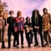 The photo shows six musicians standing next to each other on a deck near a lake. These musicians are part of the music collective Acid Mothers Temple & The Melting Paraiso U.F.O. Most of the members are uniquely dressed in colourful clothing, most probably inspired by psychedelic and retro fashion.