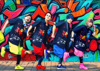 A group photo of the band Iscream Never Ground posing in front of a colourful backdrop, created by graffiti. All members are dressed in the same way, same dark shirt with paint splatter motive, but they are wearing a different colour pants and shoes. Those pants and shoes are in the same colour for each member, perhaps used as a member colour. The members stand in a row and strike a pose as if they are making an exaggerated walking motion: one leg is raised and the other is on the ground, one arm is balled into a fist and held close to their face, whilst the other arm, also with a clenched fist, is positioned behind them, with their elbows bent. © アイスクリームネバーグラウンド - Iscream Never Ground