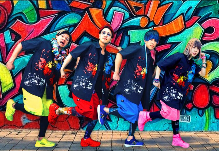 A group photo of the band Iscream Never Ground posing in front of a colourful backdrop, created by graffiti. All members are dressed in the same way, same dark shirt with paint splatter motive, but they are wearing a different colour pants and shoes. Those pants and shoes are in the same colour for each member, perhaps used as a member colour. The members stand in a row and strike a pose as if they are making an exaggerated walking motion: one leg is raised and the other is on the ground, one arm is balled into a fist and held close to their face, whilst the other arm, also with a clenched fist, is positioned behind them, with their elbows bent. © アイスクリームネバーグラウンド - Iscream Never Ground