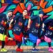 A group photo of the band Iscream Never Ground posing in front of a colourful backdrop, created by graffiti. All members are dressed in the same way, same dark shirt with paint splatter motive, but they are wearing a different colour pants and shoes. Those pants and shoes are in the same colour for each member, perhaps used as a member colour. The members stand in a row and strike a pose as if they are making an exaggerated walking motion: one leg is raised and the other is on the ground, one arm is balled into a fist and held close to their face, whilst the other arm, also with a clenched fist, is positioned behind them, with their elbows bent. © アイスクリームネバーグラウンド - Iscream Never Ground