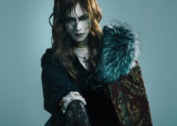 Photo of the vocalist and componist KAMIJO clothed in gothic-influenced clothing, mostly in black with lace, apart from the red with yellow/gold coat with fur-lined collar. He wears dramatic makeup, several necklaces and a white pearl armband. © CHATEAU AGENCY CO., Ltd.