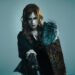 Photo of the vocalist and componist KAMIJO clothed in gothic-influenced clothing, mostly in black with lace, apart from the red with yellow/gold coat with fur-lined collar. He wears dramatic makeup, several necklaces and a white pearl armband. © CHATEAU AGENCY CO., Ltd.