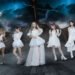 The photo features the five women of LOVEBITES posing together while standing on a ship and dark clouds with lightning on the background. They are uniformly dressed in white/off-white with gold elements outfits, each incorporating their personal style.