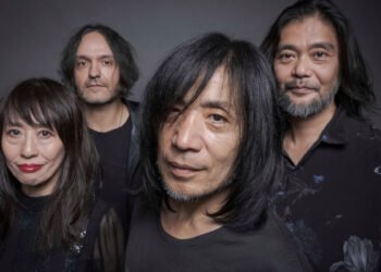 Band photo of MONO standing near each other with their faces directed to the camera lens. Some of them are smiling. The photo is dark in colour, given that all members are wearing black clothing, have dark hair, and are standing in front of a dark background, and yet it is not a black-and-white photo.