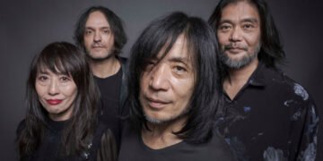 Band photo of MONO standing near each other with their faces directed to the camera lens. Some of them are smiling. The photo is dark in colour, given that all members are wearing black clothing, have dark hair, and are standing in front of a dark background, and yet it is not a black-and-white photo.