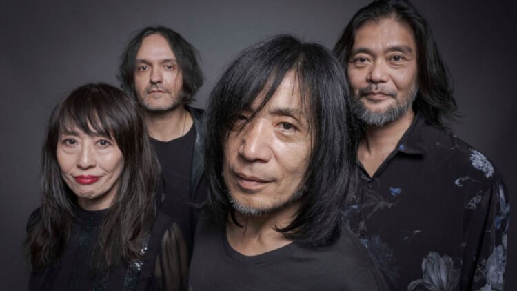 Band photo of MONO standing near each other with their faces directed to the camera lens. Some of them are smiling. The photo is dark in colour, given that all members are wearing black clothing, have dark hair, and are standing in front of a dark background, and yet it is not a black-and-white photo.