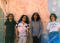 The photo features the four men of krautrock band Minami Deutsch. They are standing next to each other against a painted wall somewhere outside (in the United States), dressed casually. Most of them are wearing sunglasses as it is sunny weather. Photo by Tyler Loring @/everwavecreative on Instagram