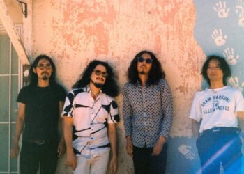 The photo features the four men of krautrock band Minami Deutsch. They are standing next to each other against a painted wall somewhere outside (in the United States), dressed casually. Most of them are wearing sunglasses as it is sunny weather. Photo by Tyler Loring @/everwavecreative on Instagram