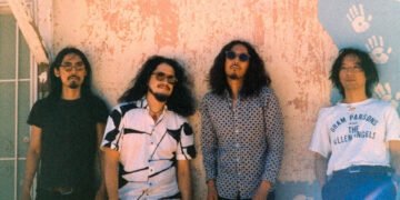 The photo features the four men of krautrock band Minami Deutsch. They are standing next to each other against a painted wall somewhere outside (in the United States), dressed casually. Most of them are wearing sunglasses as it is sunny weather. Photo by Tyler Loring @/everwavecreative on Instagram