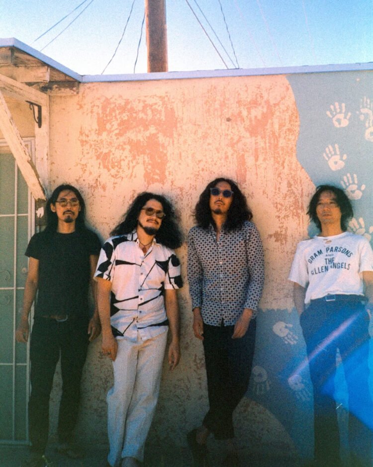 The photo features the four men of krautrock band Minami Deutsch. They are standing next to each other against a painted wall somewhere outside (in the United States), dressed casually. Most of them are wearing sunglasses as it is sunny weather. Photo by Tyler Loring @/everwavecreative on Instagram