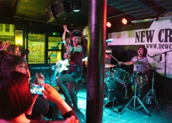 The three-piece THE LET’S GO’s in the middle of a performance at New Cross Inn in London. The guitarist and vocalist COCO is in front playing her guitar and interacting with the crowd. On the left, a bit of the audience is visible, partly enjoying the show and partly filming the show. Photography by JJ Grant (wonderlens)