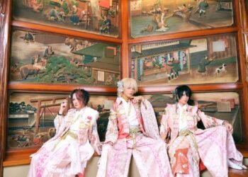 Seth, Kouki and Tsukasa dressed in traditional kimono with hakama, with pink and red hues and floral patterns. They are seated side by side with Japanese (wooden) artworks in the background.