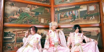 Seth, Kouki and Tsukasa of Sakura Zensen dressed in traditional kimono with hakama, with pink and red hues and floral patterns. They are seated side by side with Japanese (wooden) artworks in the background. © Sakura Zensen