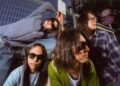 Band photo of the four members of DYGL posing in front of a camera, but most are not looking into the lens and two of them are even looking away. The musicians are dressed casually and all wearing sun glasses. They are posing in front of a gate and a train is visible in the background. Photography: Earl Standerford
