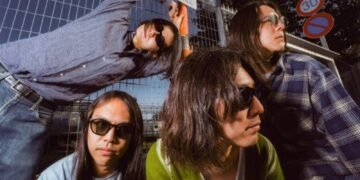 Band photo of the four members of DYGL posing in front of a camera, but most are not looking into the lens and two of them are even looking away. The musicians are dressed casually and all wearing sun glasses. They are posing in front of a gate and a train is visible in the background. Photography: Earl Standerford